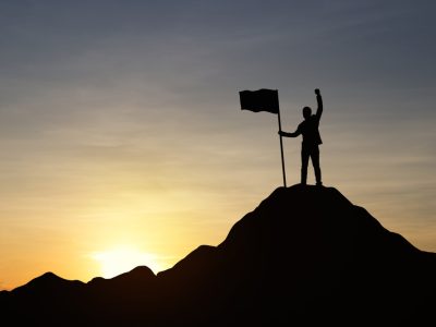 Silhouette,Of,Businessman,Holding,A,Flag,On,Top,Mountain,,Sky